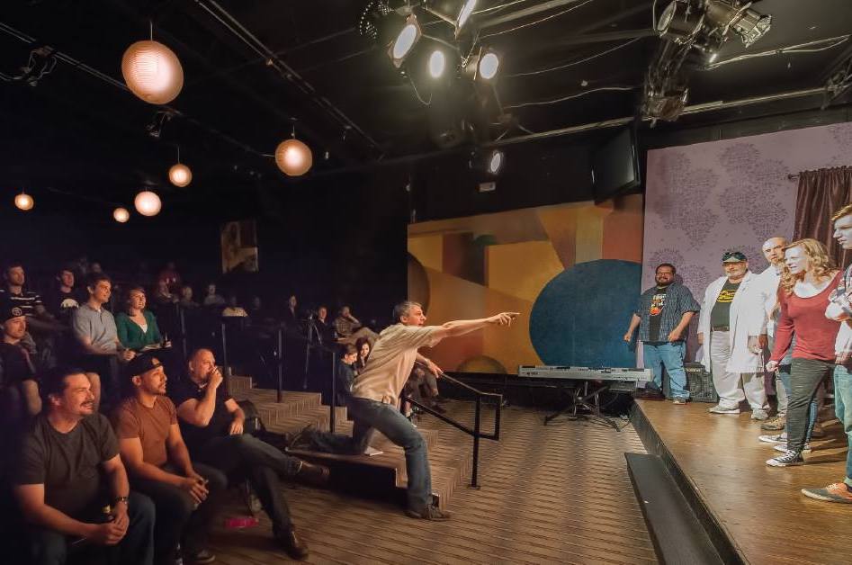 The Science Improv event in 2022. Smiling audience members can be seen looking at the stage where performers are looking attentively at Lew Lefton. Lew points to the performers, giving an animated explanation of the next improv prompt. 