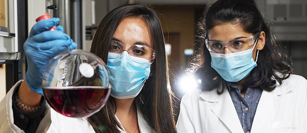Graduate students Kirstie Thompson (left) and Ronita Mathias with a flask containing polymer materials that are being used to create a new membrane technology — one that could reduce carbon emissions and energy intensity associated with refining crude oil. (Credit: Christopher Moore)